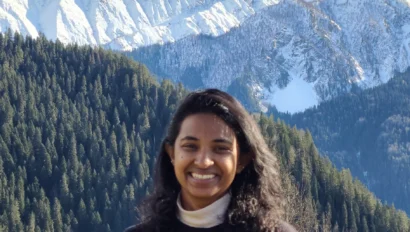 A person smiling in front of snow-covered mountains and pine forests under a clear blue sky.
