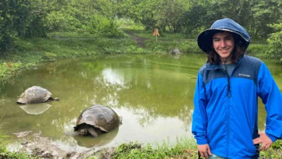 A person in a blue rain jacket and boots stands by a pond with two large tortoises, surrounded by lush greenery on a conservation trip.
