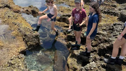 A group of people explore a rocky tide pool by the ocean, wearing shorts and boots.