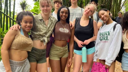 A group of smiling young adults in casual summer clothing stand together outdoors in a bamboo grove.