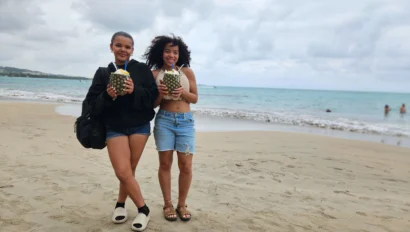 Two women standing on a beach holding pineapples with drinks inside, with people and ocean waves in the background.