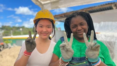 Two people wearing work gloves and smiling while holding up peace signs at a construction site. One is wearing a hard hat.