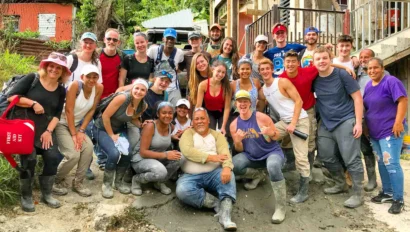 A group of students, smiling and wearing casual clothes and boots, pose together outdoors in front of a partially constructed building service project during Global Works' student travel to Puerto Rico.