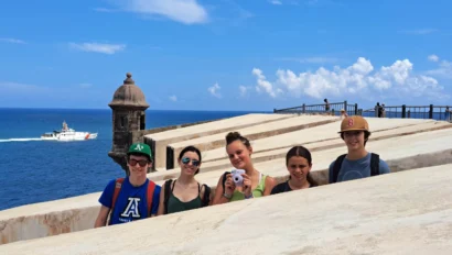 Five people stand on a stone structure near the ocean, with a boat in the background under a blue sky.