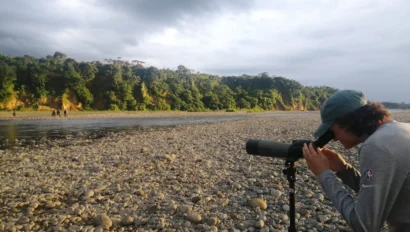 Person using a telescope on a rocky riverbank with a forest in the background under a cloudy sky.
