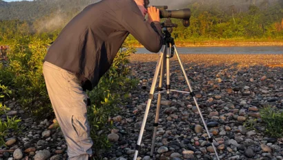 Person in boots using a telescope on a tripod, standing on rocky terrain with a mountain backdrop.