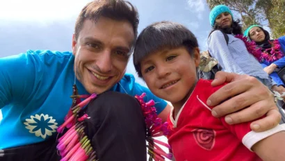A man in a blue shirt and a boy in a red shirt smile at the camera. They wear colorful necklaces. Two people in the background are also visible under a clear sky.