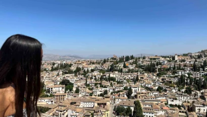 Woman looking out over a sprawling cityscape with terracotta rooftops, trees, and distant mountains under a clear blue sky.