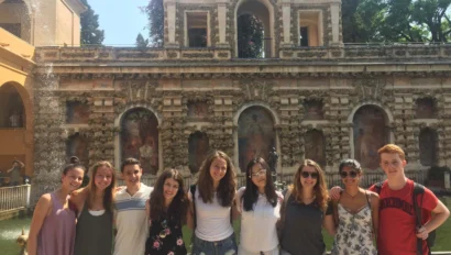 Group of nine people standing in front of an ornate building with arches and textured walls, posing for a photo.