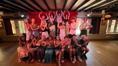A group of people posing indoors, some standing and making gestures while others are seated, in a warmly lit room with wooden beams and red curtains.