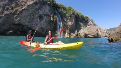 Two people kayaking on blue water in front of a rocky cliff with a waterfall under a clear sky.