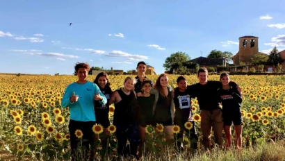 A group of nine people stands together in a sunflower field under a clear sky, enjoying a break from their Spanish immersion program, with a building and trees visible in the background.