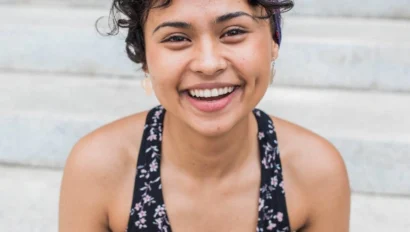 A person with curly hair and a headband smiles while sitting on outdoor steps, wearing a floral-patterned top.