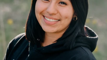 A person with shoulder-length black hair smiles while wearing a black hoodie with a logo, standing outdoors with a blurred green background.
