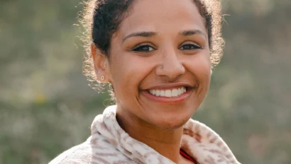 A person with curly hair in a bun smiles outdoors, wearing a light patterned jacket.