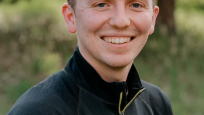 A person in a black jacket smiles outdoors with greenery in the background.