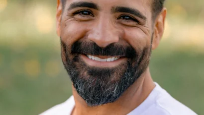 Man with a beard and tied back hair smiling outdoors, wearing a white shirt.