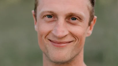 A smiling man with short blond hair wearing a light blue shirt stands outdoors with a blurred green background.