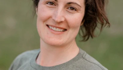 A person with a ponytail smiles at the camera, wearing a gray T-shirt with orange lettering and a footprint design.