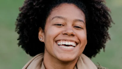 Smiling person with curly hair wearing a tan scarf and a green fleece jacket, standing outdoors.