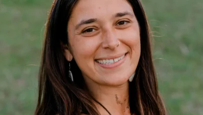 Person with long brown hair smiling outdoors, wearing a tie-dye shirt.
