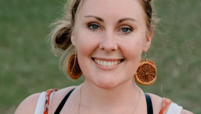 A woman with light hair and orange attire smiles outdoors, wearing large circular earrings.