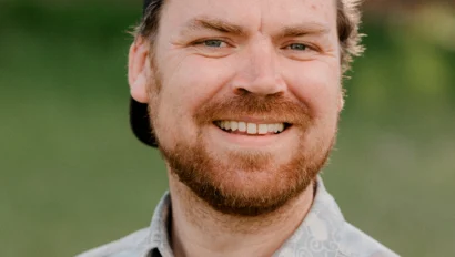 A man with a beard wearing a backward cap and patterned shirt smiles outdoors with a blurred green background.