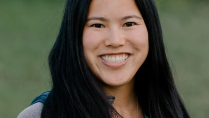 A smiling person with long dark hair wearing a blue vest and gray top stands outdoors.