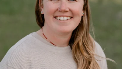 A person with long hair is smiling outdoors, wearing a light sweater. The background is a grassy area.