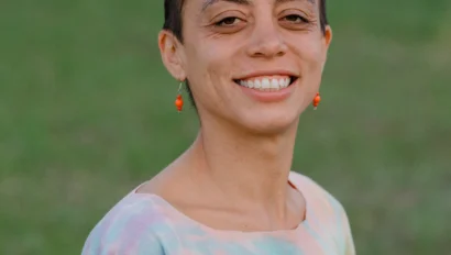 Person with short hair smiling, wearing a tie-dye shirt and orange earrings, standing outdoors on grass.