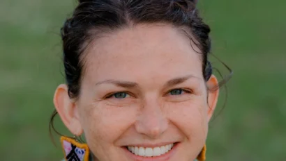 A person with brown hair in a bun, wearing a green hoodie and colorful earrings, smiles in an outdoor setting.