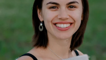 A person with short brown hair smiles at the camera, wearing a light-colored off-shoulder sweater, earrings, and a necklace, standing in front of a blurred green background.