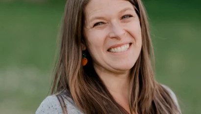 A woman with long brown hair smiles at the camera, standing outdoors with a grassy background. She is wearing a gray sweater over a green top.