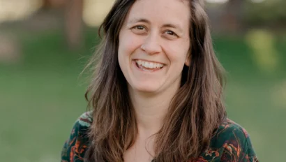 A person with long brown hair, wearing a floral-patterned shirt, smiles outdoors in a grassy area.