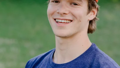 A person wearing a blue shirt and red cap smiles outdoors with blurred greenery in the background.