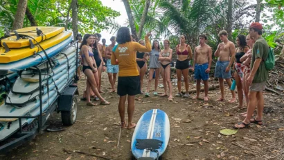 A group of people stand in a circle on a beach, listening to an instructor near a stack of surfboards and a single board on the ground.