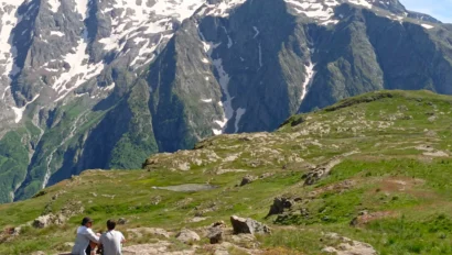 Two people sit on a grassy hill overlooking a mountain range with snow-capped peaks under a clear blue sky.