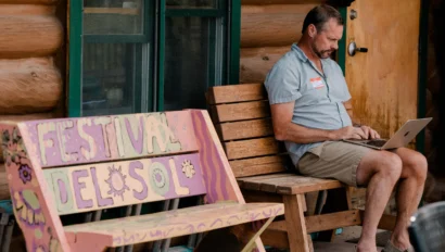 Man sitting on a wooden bench working on a laptop outside a log cabin. A colorful sign reading "Festival del Sol" is beside him.