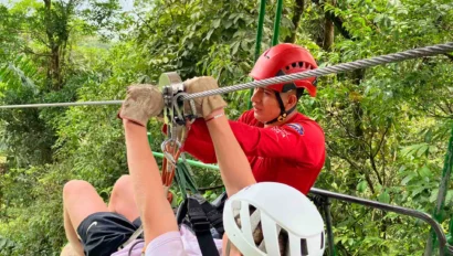 Person in white helmet preparing for a zipline ride with the assistance of an instructor in a red helmet, surrounded by lush green forest.