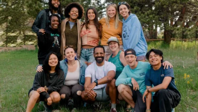 A diverse group of twelve people smiling and posing outdoors on a grassy field with trees in the background.