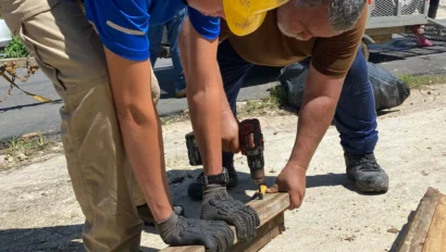 Two people in work gear use power drills to attach wooden beams on a sunny day.
