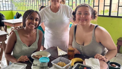 Two people sitting at a table with food containers and bananas, while an older person stands behind them. They are in a brightly lit room with green walls and a windowed background.