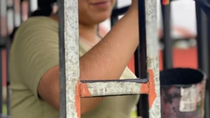 A person paints a metal fence with orange paint using a brush, holding a can of paint in the other hand.