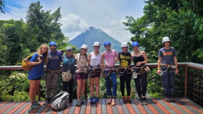 A group of people wearing helmets and harnesses stand on a wooden deck with trees and a mountain in the background.