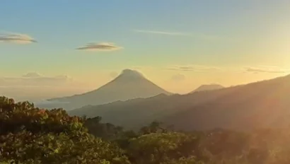 Panoramic view of a sunrise over a landscape with a distant mountain, surrounded by trees and rolling hills.