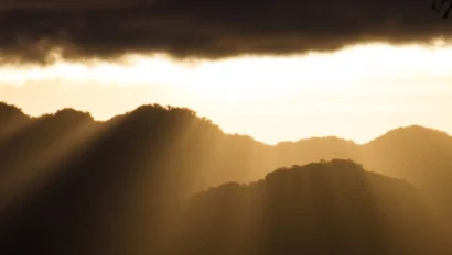 Silhouetted mountains with sun rays breaking through clouds, framed by palm trees.
