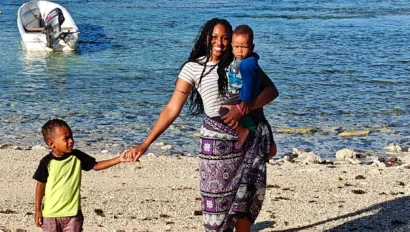 A woman stands on a beach during a Fiji school trip, holding a small child and the hand of another, with a boat and mountains in the background.