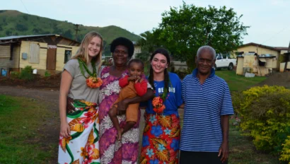 Five people, including a child, stand together outside in a village setting during a Fiji school trip, smiling at the camera. Two women wear leis and colorful skirts, with houses and greenery in the background.