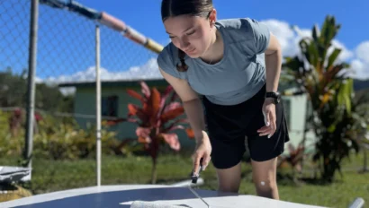 A person in a grey shirt and black shorts uses a paint roller to apply white paint outdoors on a sunny day, helping prepare for the upcoming Fiji school trip.