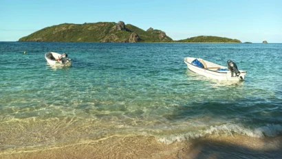 Two small motorboats float near the shore in clear, shallow water during a Fiji school trip, with an island and blue sky in the background.